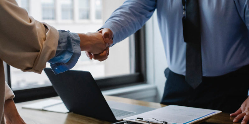 Business Man Behind Desk with a Laptop and Documents Shaking a Business Woman's hand