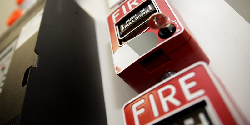 Two fire alarms on top of each other near a electrical box on a wall